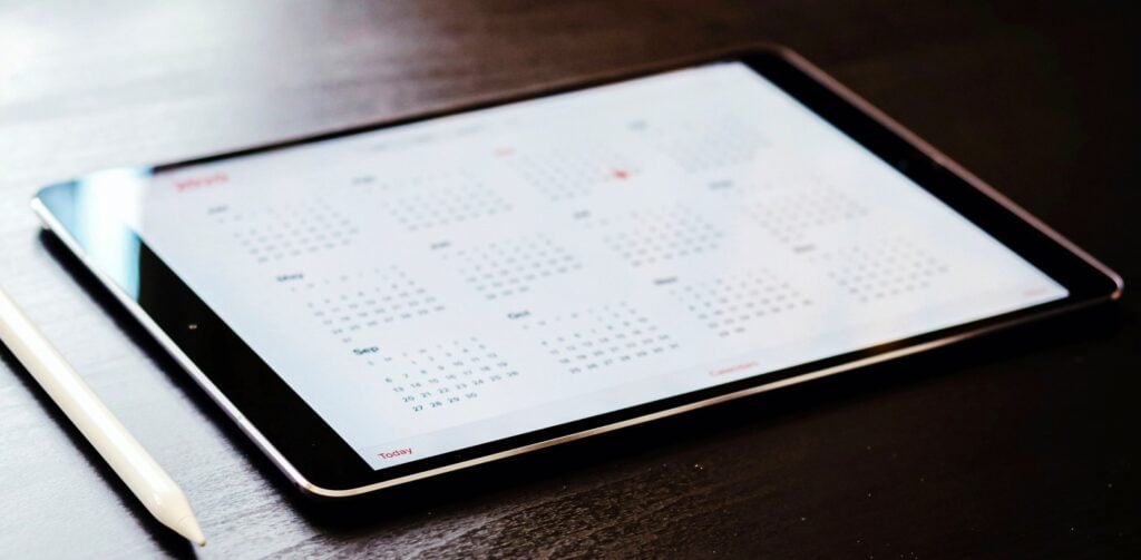 A tablet on a mahogany desk with a calendar app open and a white stylus pen on the left.