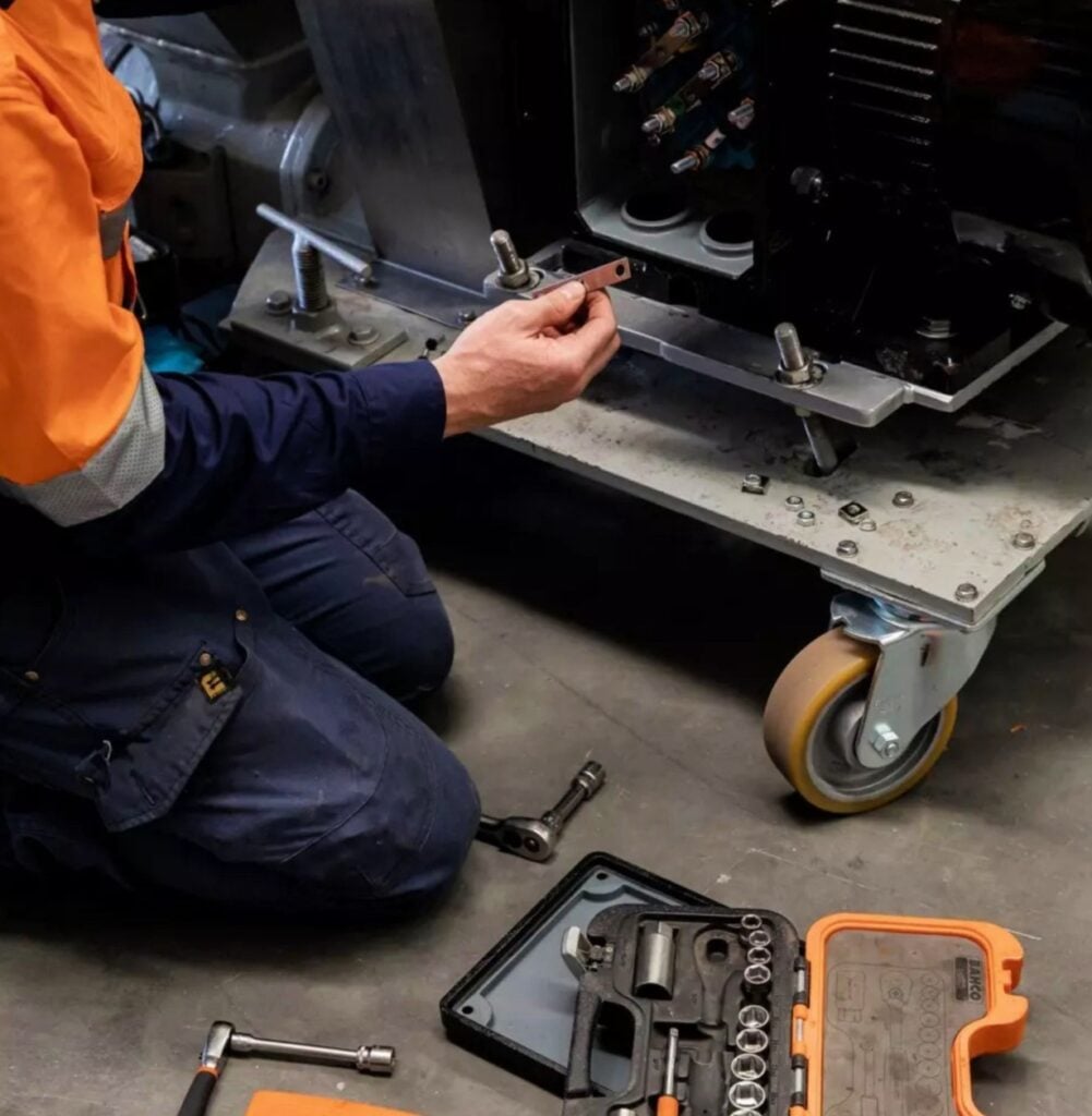 A maintenance technician in dark blue and orange overalls with an open toolbox full a different sized wrench heads repairing a machine.