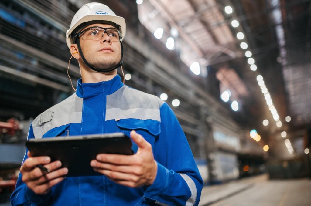 A maintenance technician wearing blue overalls, a hard hat, and safety goggles holding a tablet.
